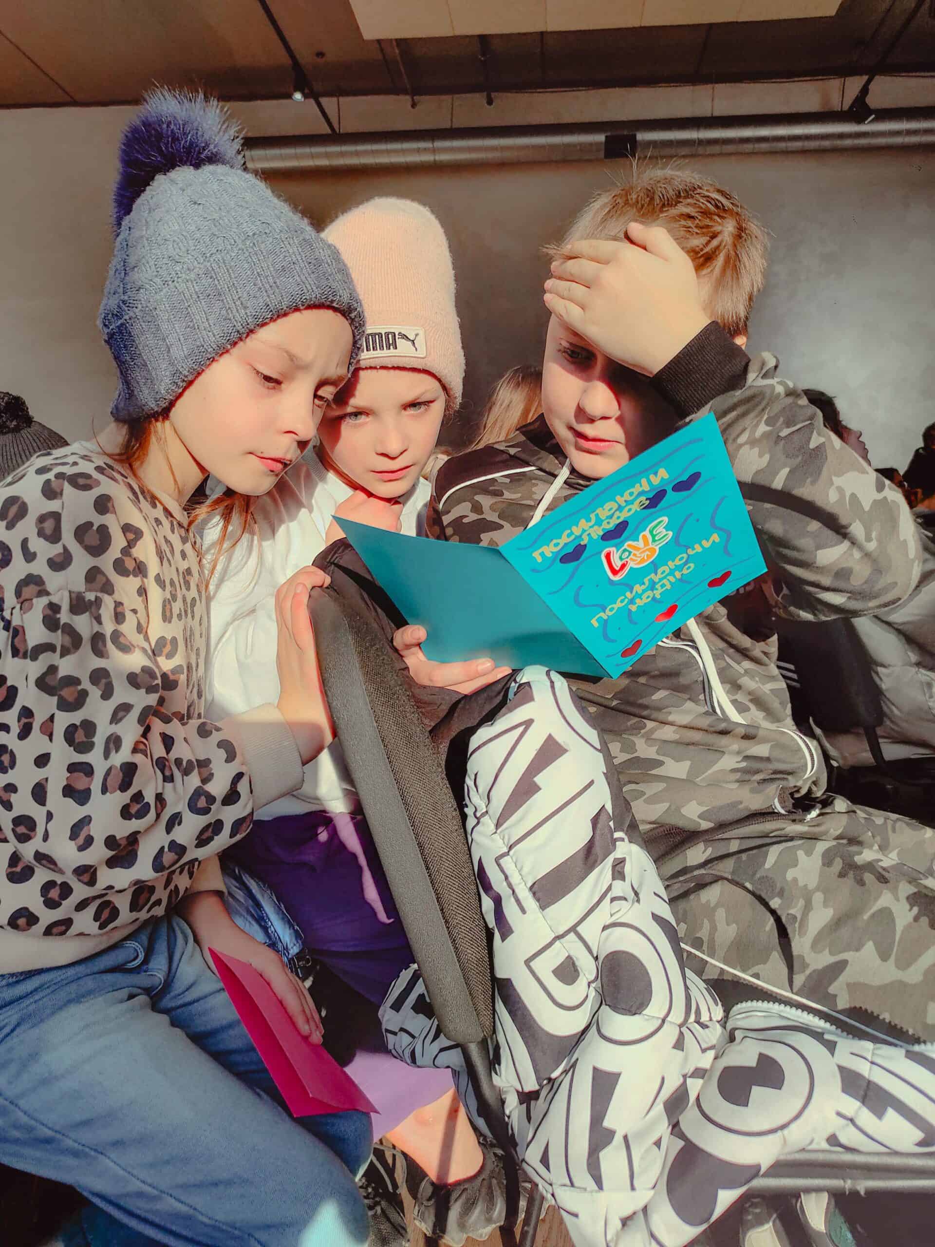 Three young Ukrainian children huddle together, reading a bright blue letter with curiosity and emotion. Sunlight streams in, highlighting their faces as they take in the heartfelt message.