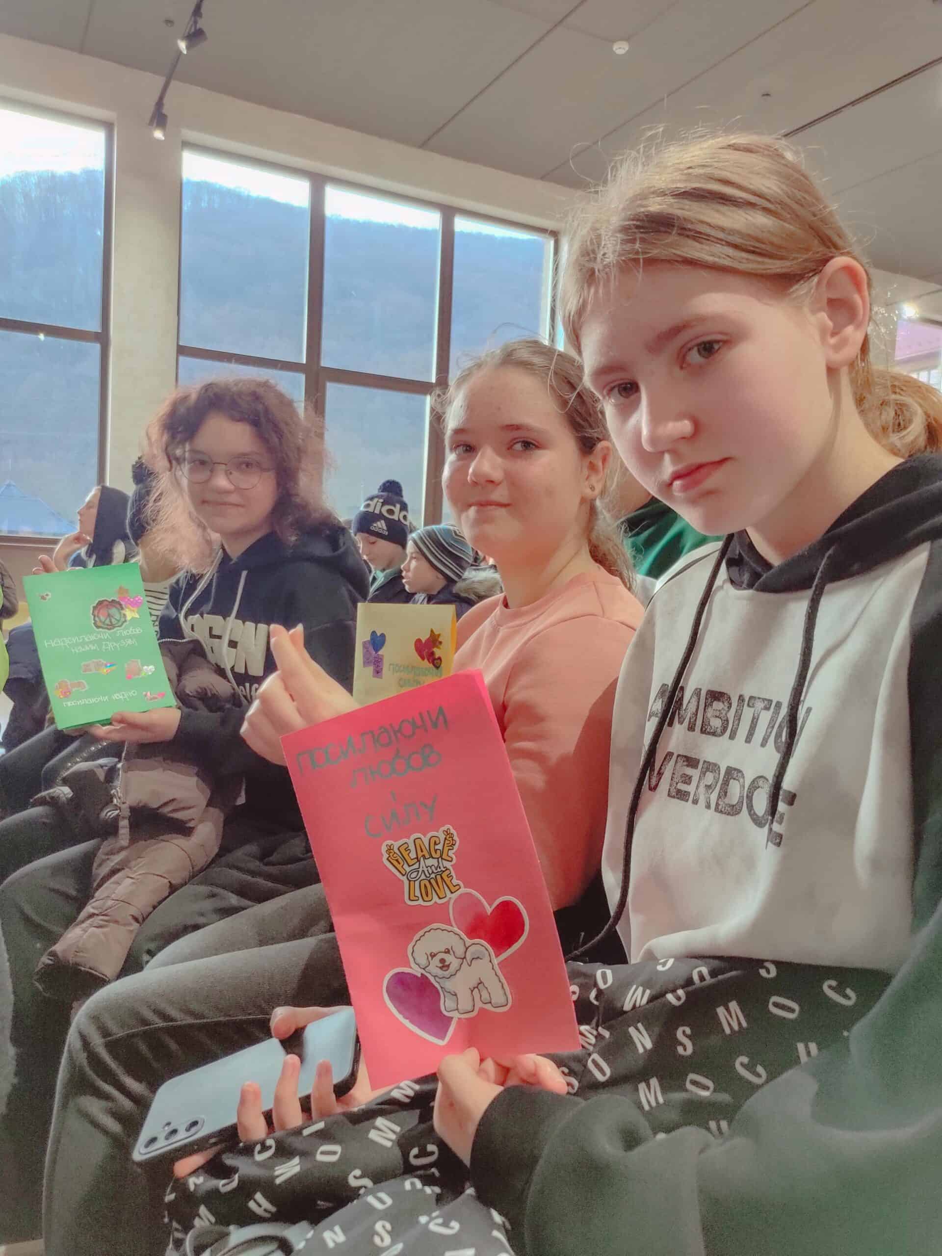 Three Ukrainian girls sit together, holding handmade letters decorated with stickers and bright colors. They smile gently as they share messages from American students, with a mountain landscape visible through the windows behind them.