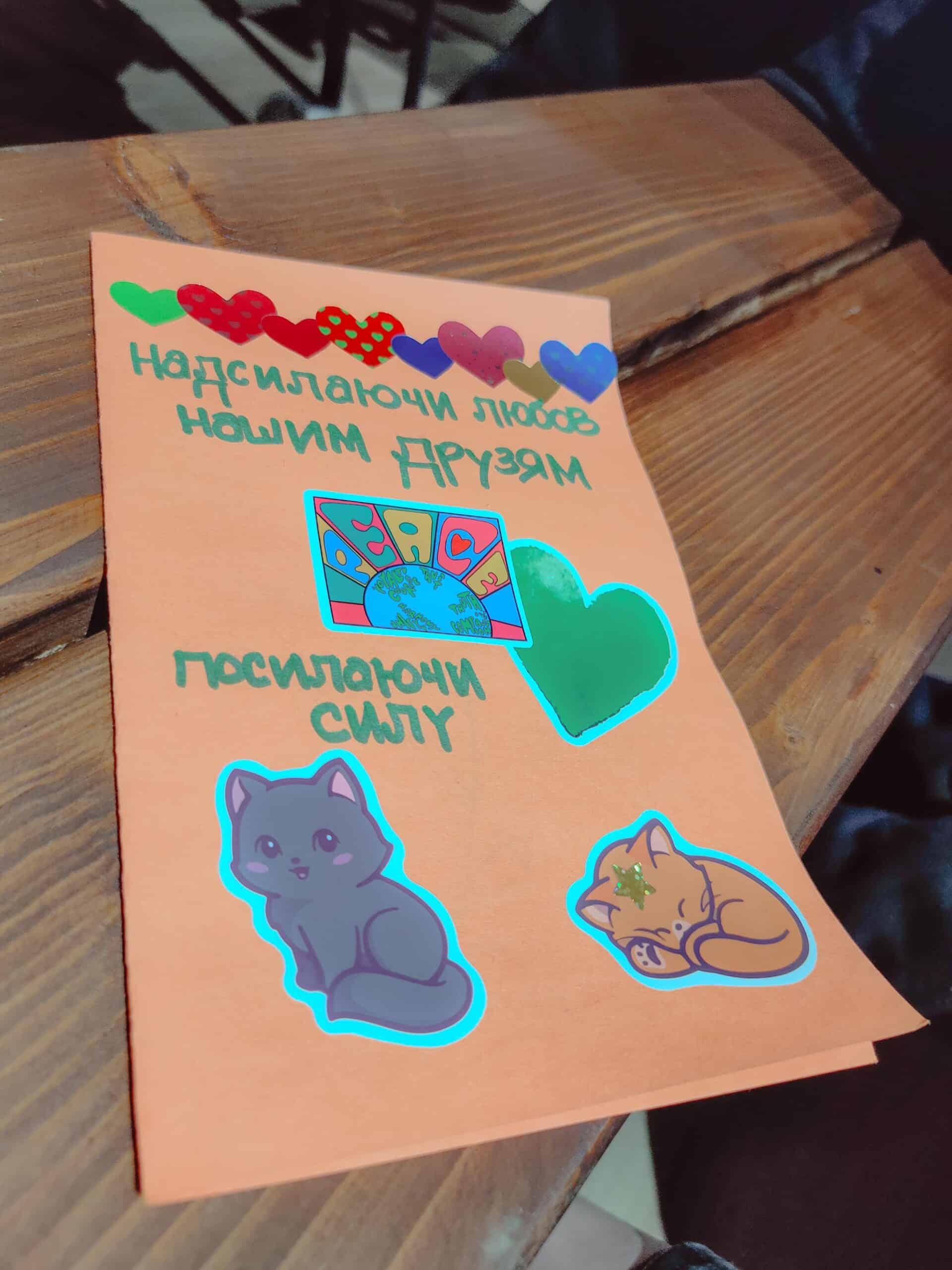 A handmade orange card with Ukrainian text, colorful heart stickers, a "Peace" emblem, and two cute cat stickers sits on a wooden table. The message sends love and strength to new friends.