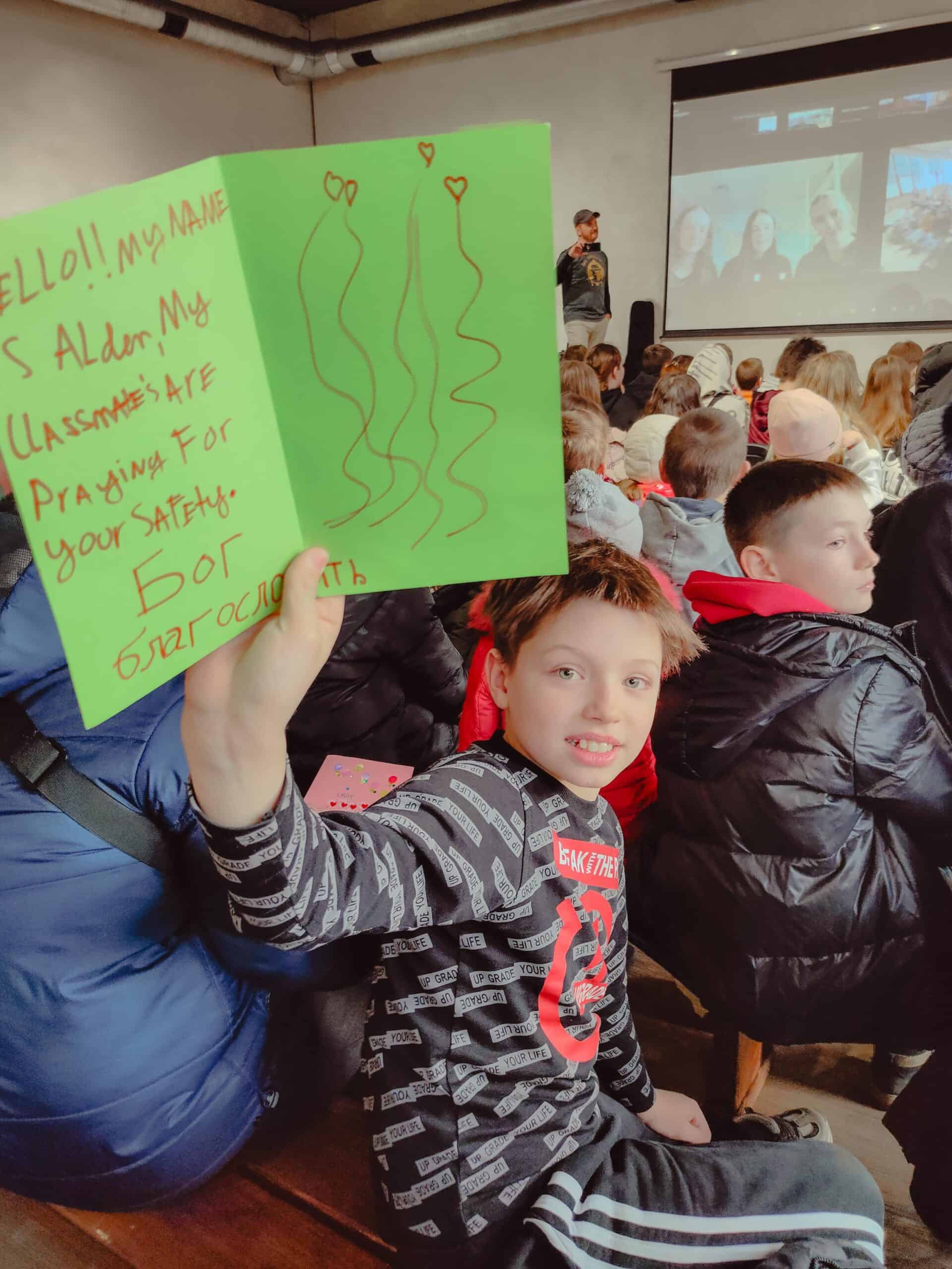 A young Ukrainian boy, smiling, holds up a bright green card with an English message: "HELLO! My name is Alder. My classmates are praying for your safety." The background shows a large gathering of children watching a virtual meeting.