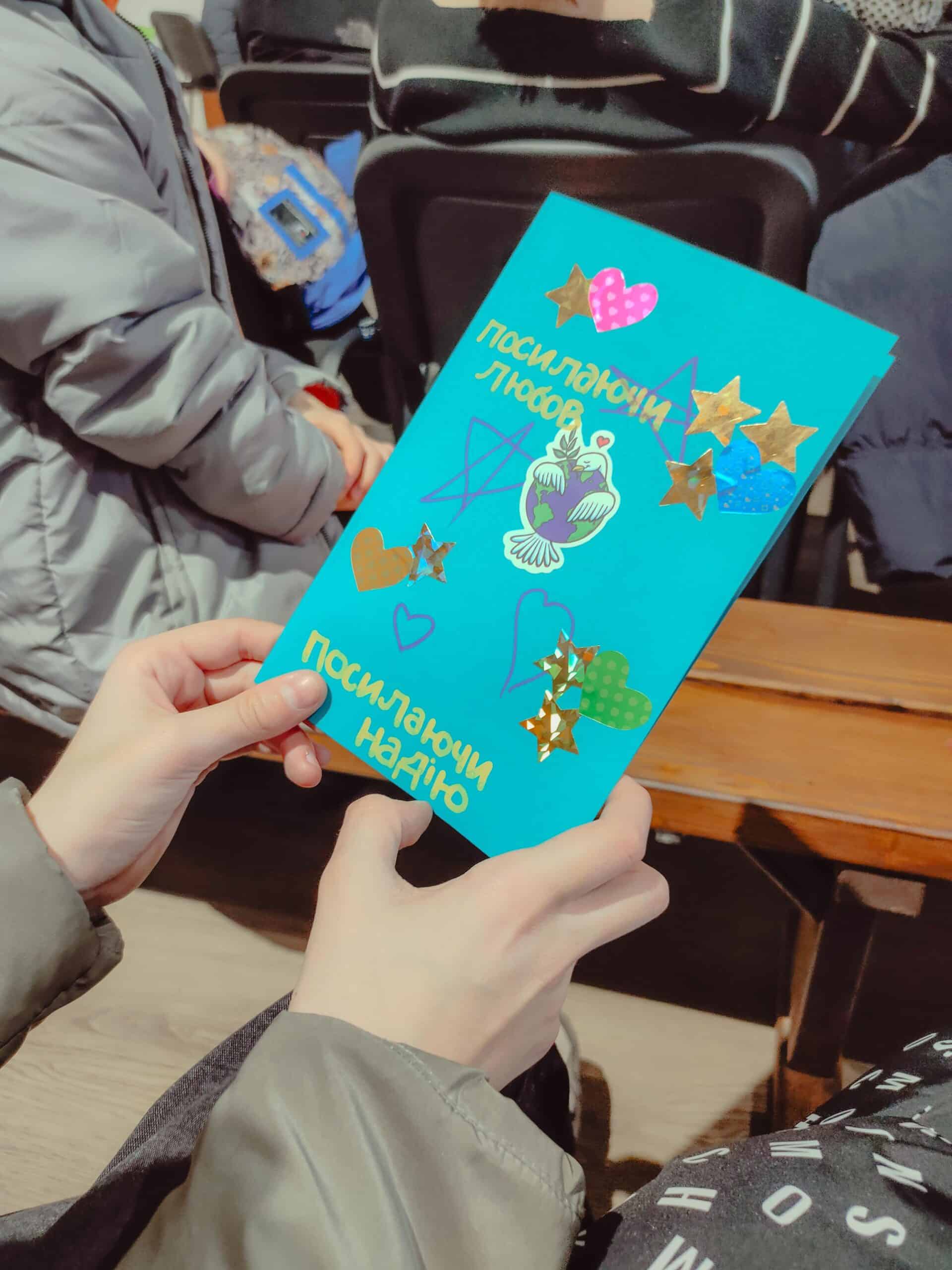 A close-up of a bright blue handmade card with Ukrainian text reading "Sending Love" and "Sending Hope," decorated with shiny gold stars, colorful hearts, and a peace dove sticker.
