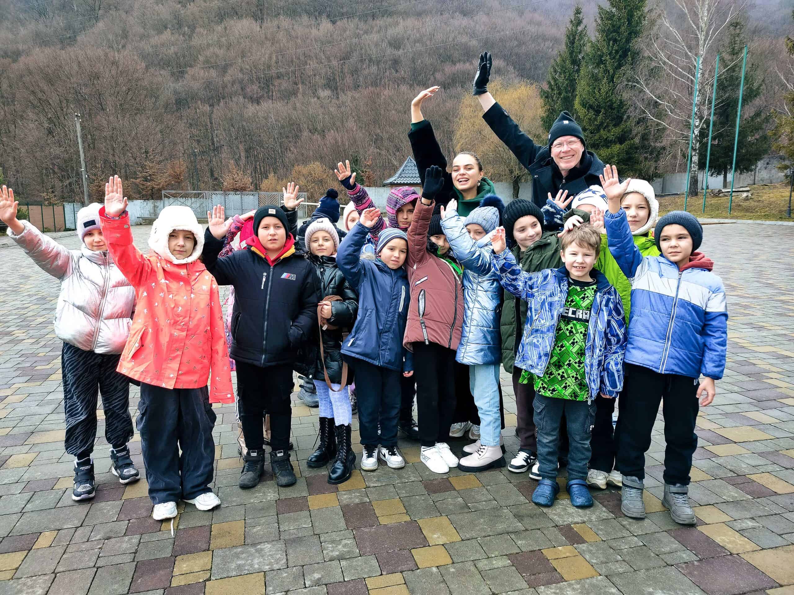 FH - CampDreamlandWinter-311 Group of Ukrainian children and two adults enthusiastically raising their hands in a courtyard surrounded by forested hills.
