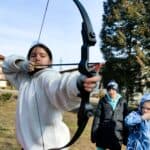 A Ukrainian girl in a white fleece jacket holds a bow and arrow, concentrating as she aims.
