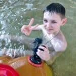 A smiling boy with wet hair gives a peace sign while sitting in the shallow pool.