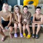 Four kids in swimsuits and towels sit on lounge chairs by the pool, drying off after swimming.