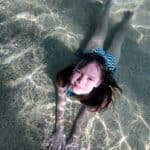 A Ukrainian girl in a striped swimsuit floats on her stomach in a sunlit pool, smiling up at the camera.