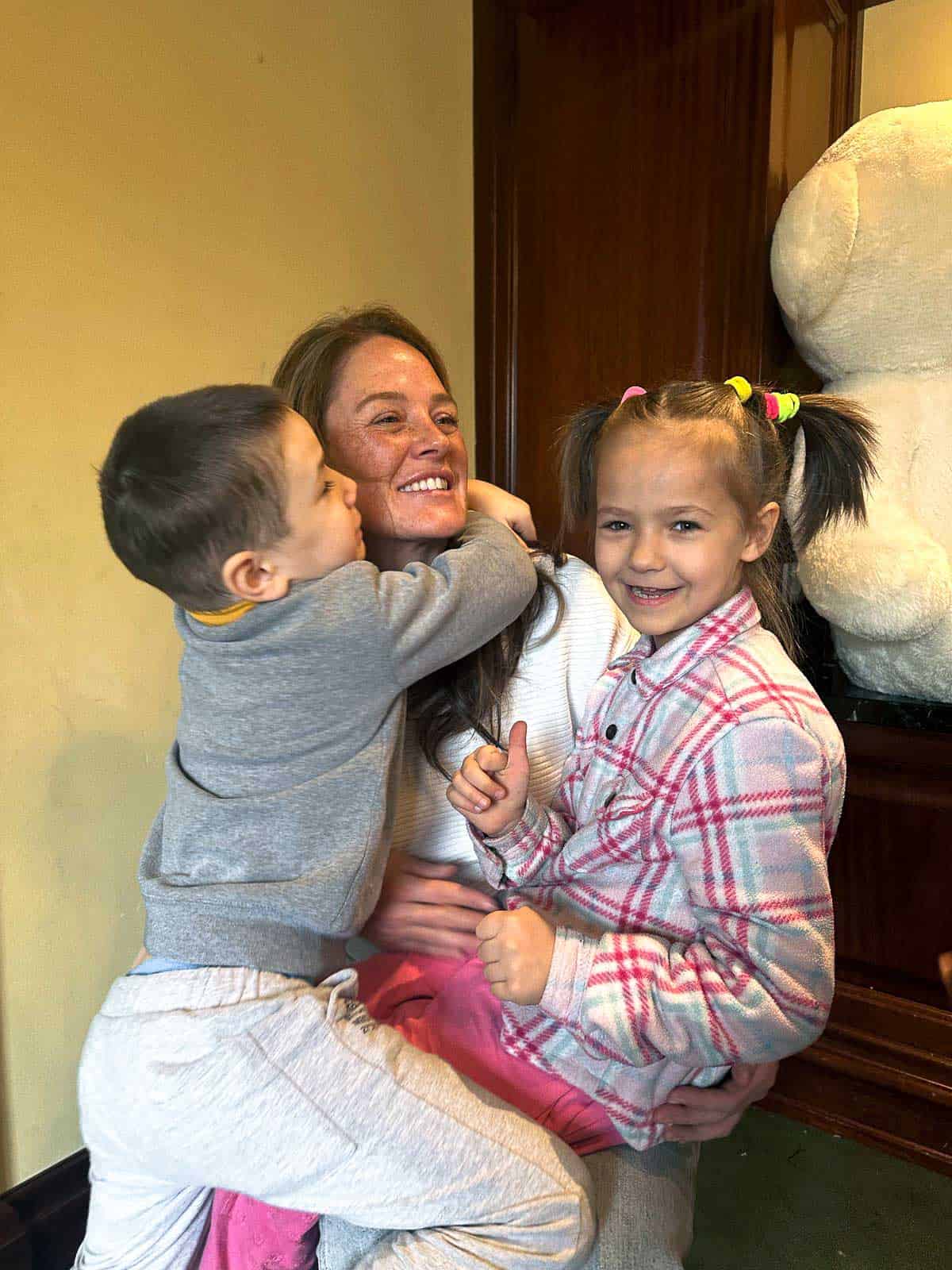 A woman holds two young children, one hugging her and the other grinning while showing a thumbs-up.