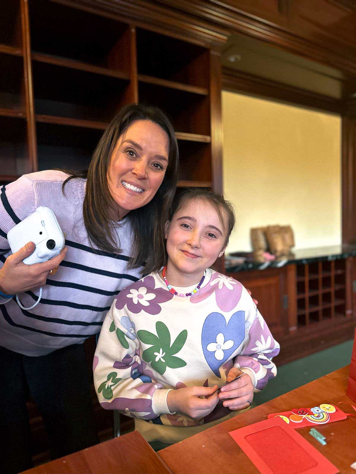 A young girl in a colorful sweater sits at a table with an adult volunteer, working on a craft project together.