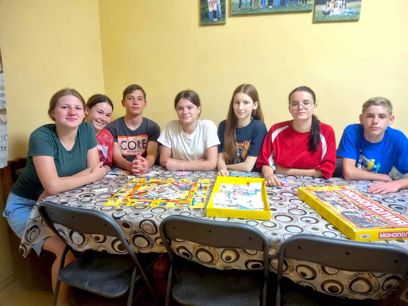 Seven Ukrainian foster children sit together at a dining table in their home, smiling and gathered around a Monopoly board game.