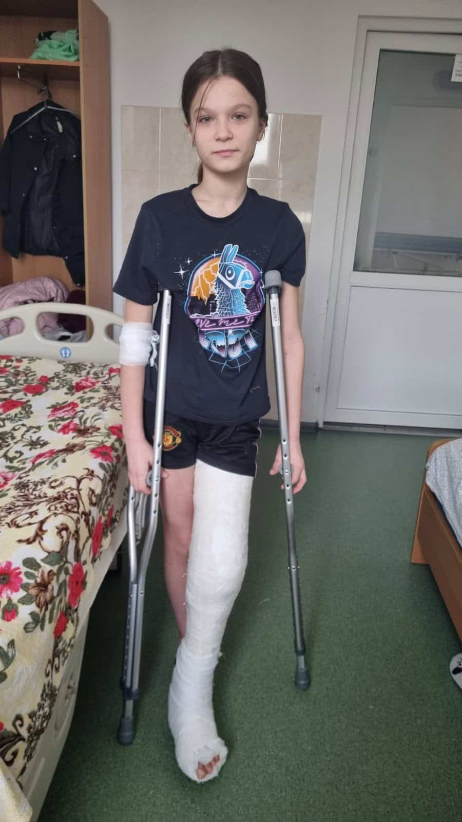 Ukrainian foster child Veronika stands with crutches in a hospital room, her leg in a full cast after surgery.