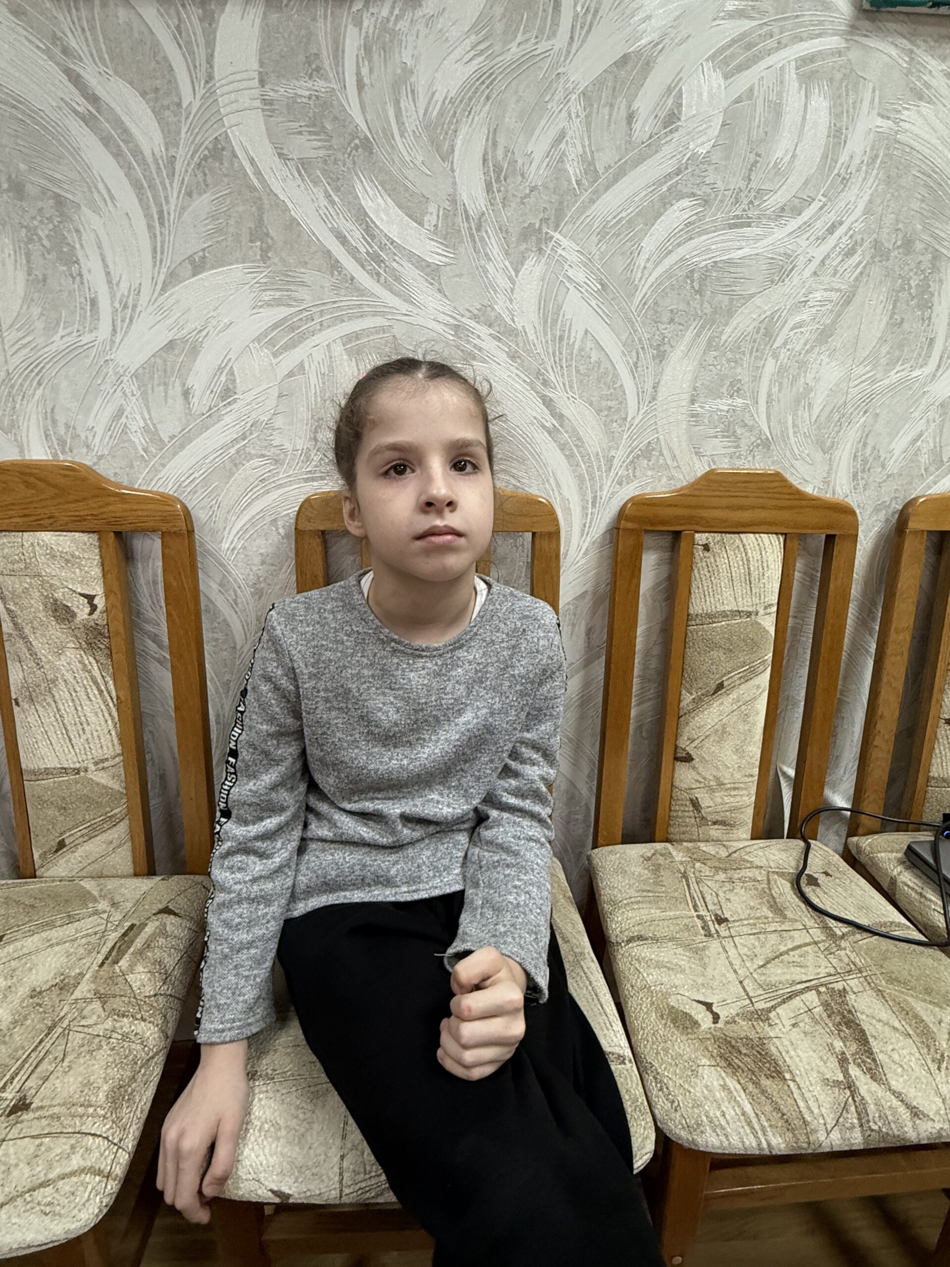 A quiet, sweet 9-year-old Ukrainian girl named Anastasia, looking softly at the camera with a gentle expression. She is photographed against a simple, neutral background.